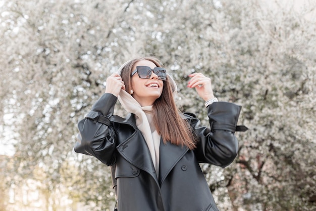 Gelukkig mooie jonge hipster vrouw met positieve emoties in modieuze casual kleding met leren jas en hoodie met trendy zonnebril wandelen en genieten in een bloeiend lentepark