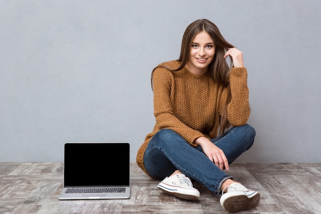 Gelukkig mooie jonge vrouw in bruine trui, jeans en sneakers zittend op houten vloer in de buurt van laptop