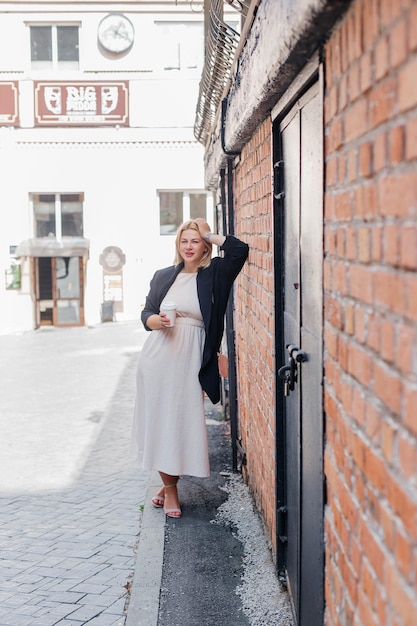 Foto gelukkig mooie jonge vrouw in een jurk en jas staat bij een bakstenen muur en drinkt koffie