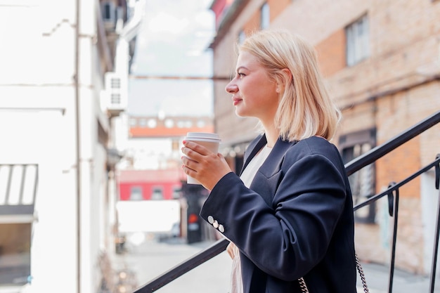 Gelukkig mooie jonge vrouw in jurk en jas loopt door de stad en drinkt koffie