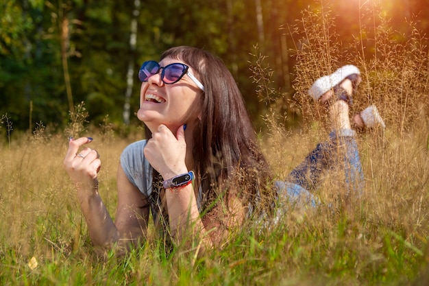 Gelukkig mooie jonge vrouw in zonnebril glimlacht en ligt op een weide in het gras in de zon