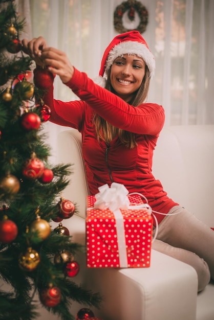 Gelukkig mooie jonge vrouw kerstboom versieren met rode sieraad thuis.