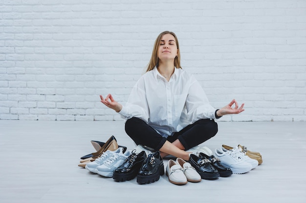 Gelukkig mooie jonge vrouw met een glimlach in een modieuze outfit met een wit overhemd en zwarte broek zit op de vloer tussen de schoenen en kiest een nieuw paar schoenen voor zichzelf