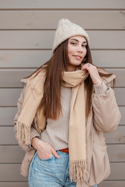 Gelukkig mooie jonge vrouw met een glimlach in modieuze winterkleren met een gebreide muts, jas, trui, sjaal en spijkerbroek in de buurt van een houten muur