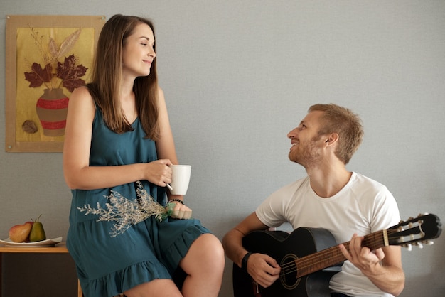 Gelukkig mooie jonge vrouw met kopje koffie kijken naar lachende vriendje gitaar spelen en ondertekenen
