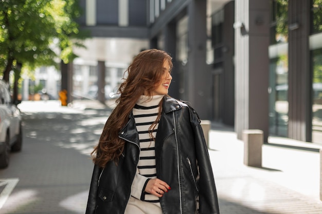Gelukkig mooie modieuze vrouw met een glimlach in stijlvolle casual kleding met een leren jas loopt op een zonnige dag in de stad