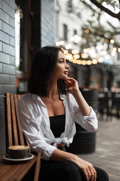 Gelukkig mooie vrouw in modieuze casual kleding met een wit overhemd zit in een zomercafé in de buurt van een zwarte bakstenen muur en drinkt koffie tegen een achtergrond van bokeh lichten