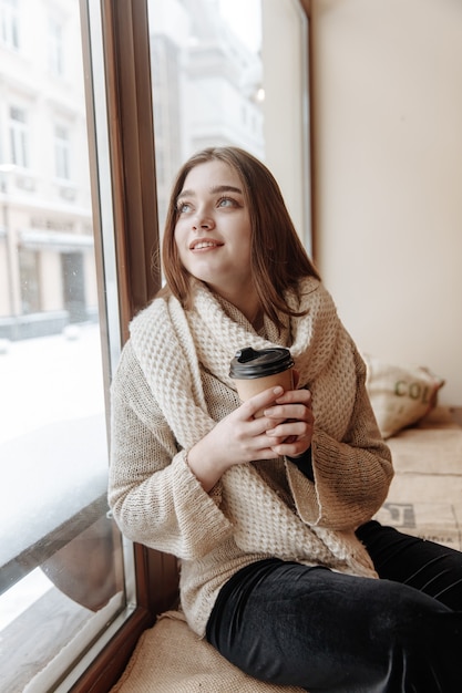 Gelukkig mooie vrouw in witte trui en sjaal zitten in de buurt van venster in café