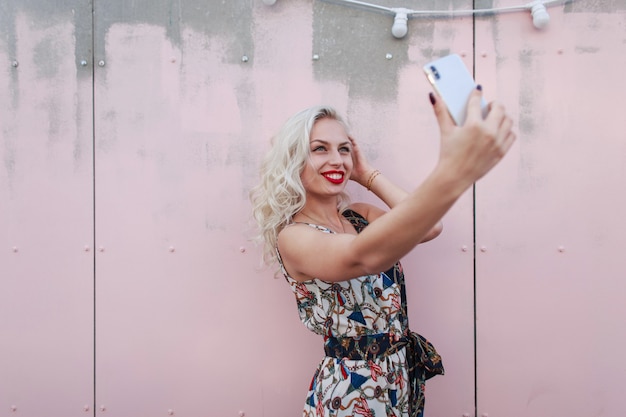 Gelukkig mooie vrouw met een glimlach in een vintage jurk selfie op een smartphone in de buurt van een roze muur op straat