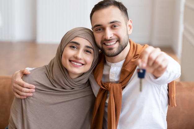 Gelukkig moslim familie paar tonen nieuwe huissleutel aan Camera zittend op de Bank binnenshuis. Eigen huis, eigendom van onroerend goed en huisvesting. Hypotheek en flataankoop. Selectieve focus