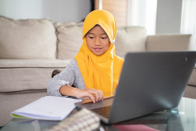 Gelukkig moslimkind dat een hoofddoek draagt die lacht terwijl hij thuis een laptop gebruikt