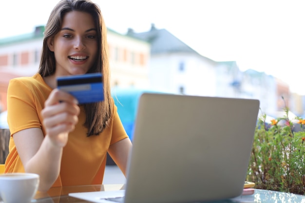 Foto gelukkig natuurlijke brunette met creditcard en laptop in café buitenshuis.