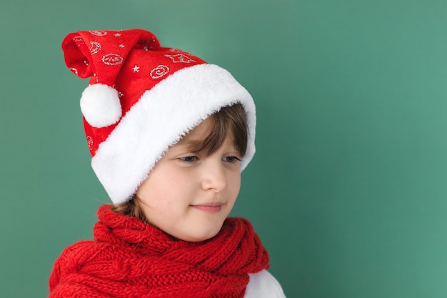 Gelukkig nieuwjaar en vrolijk kerstfeest. schattig klein kaukasisch meisje in een kerstmuts en een rode sjaal op een groene muur met plaats voor tekst.