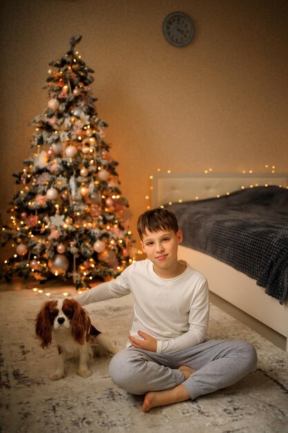 Gelukkig nieuwjaar knuffelt een jongen in lichte zelfgemaakte pyjama's zijn huisdier cavalier king charles spaniël thuis in de slaapkamer bij de kerstboom