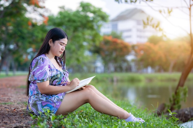 Gelukkig ontspannen portret van freelancer Aziatische vrouw Draag paarse jurk tijdens het werken met dagboek boek schrijven notitie zittend op groen gras gazon naast een reservoir in het stadspark buitenshuis