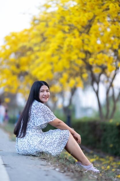 Gelukkig ontspannend portret aziatische vrouw in mooie bloeiende gele gouden tabebuia chrysotricha bloemen met het park in lentedag bij avondachtergrond in thailand