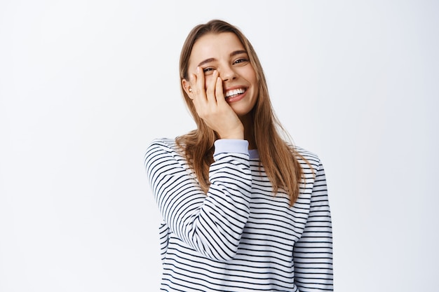 Gelukkig openhartig meisje met natuurlijke lichte make-up, gezicht aanraken en lachend met witte gezonde tanden, zorgeloos tegen de witte muur staan