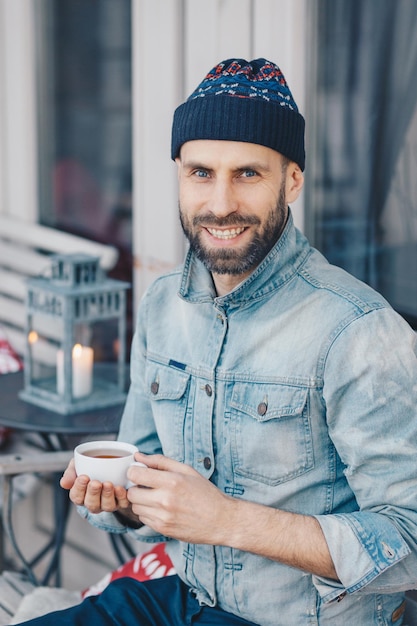 Gelukkig opgetogen bebaarde man van middelbare leeftijd met positieve uitdrukking draagt een stijlvolle hoed en denim shirt drinkt hete thee in een goed humeur geniet van vrije tijd mensen vrije tijd en recreatie concept