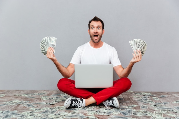 Gelukkig opgewonden casual man zittend op de vloer met laptop en geld aanhouden over grijze achtergrond