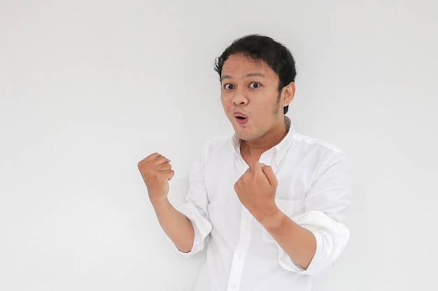 Gelukkig opgewonden en glimlachende jonge Aziatische man die zijn arm opsteekt om succes of prestatie te vieren Indonesische man met wit overhemd