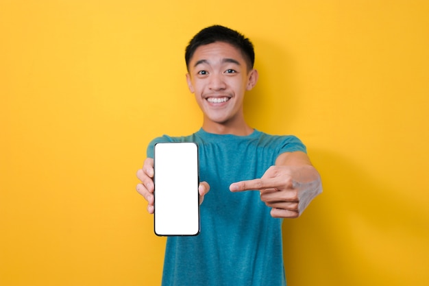 Gelukkig opgewonden jonge Aziatische man schok wijzend op leeg telefoonscherm voor kopie ruimte en toont wit telefoonscherm op camera, geïsoleerd op gele achtergrond