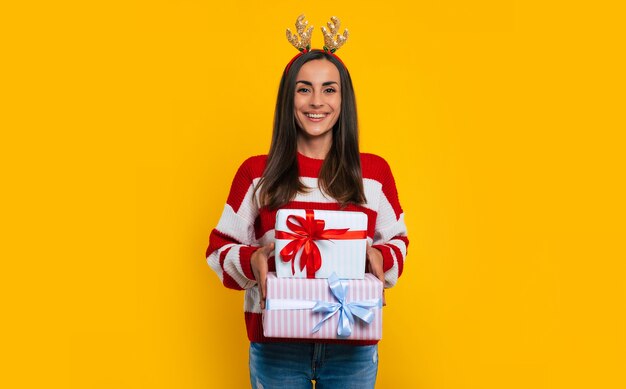 Gelukkig opgewonden jonge brunette vrouw in herten hoed, met veel kleurrijke kerst geschenkdozen in handen geïsoleerd op gele achtergrond.