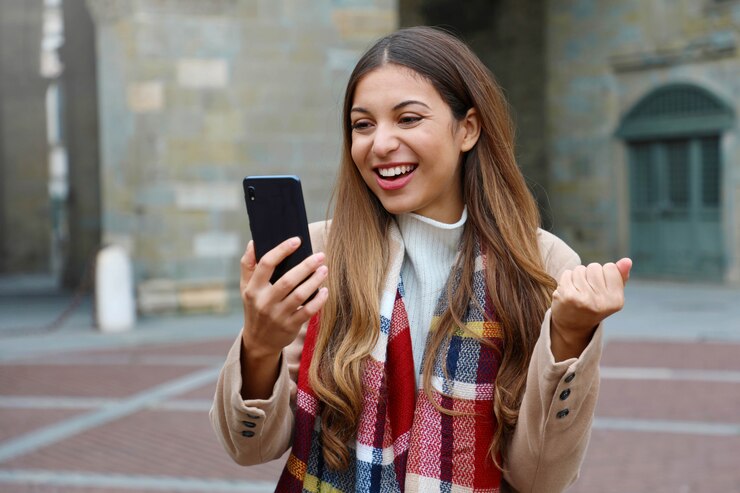 Gelukkig opgewonden jonge vrouw lacht kijken naar goed nieuws op mobiele telefoon met vuist omhoog in stad straat, wintertijd