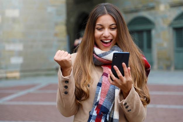 Gelukkig opgewonden jonge vrouw lacht kijken naar goed nieuws op mobiele telefoon met vuist omhoog in stad straat, wintertijd