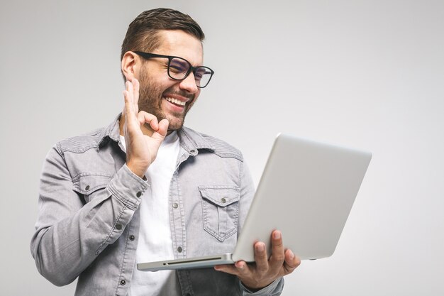 Gelukkig opgewonden laptop van de jonge mensenholding en het opheffen van zijn wapen om succes of verwezenlijking te vieren. Ok teken. Witte achtergrond.