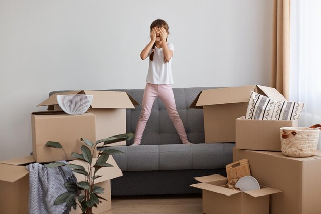 Gelukkig opgewonden meisje met vlechten, gekleed in casual stijl kleding, staand op de bank omringd met dozen met persoonlijke stapel, ogen bedekken met handpalmen, pee-a-boo spelen, bewegen vieren.