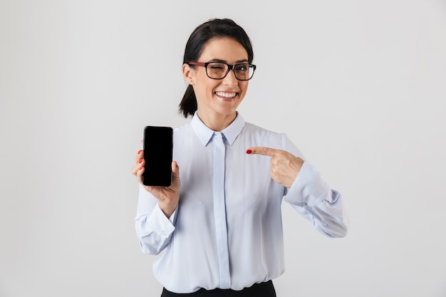 Gelukkig opgewonden zakenvrouw netjes gekleed staande geïsoleerd over witte muur, wijzend op lege mobiele telefoon