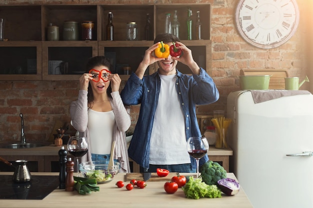 Gelukkig paar dat gezond voedsel samen kookt in hun loftkeuken thuis. Man en vrouw grimassen met verse peper, vasthouden als een bril tijdens het bereiden van groentesalade, kopiëren ruimte