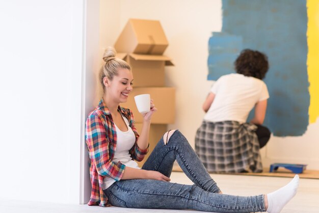 Foto gelukkig paar dat huisrenovatie doet, de man schildert de kamer en de vrouw ontspant op de vloer en drinkt koffie