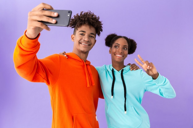 Gelukkig paar dat kleurrijke sweatshirts draagt die selfie op smartphone nemen, die over violette muur wordt geïsoleerd