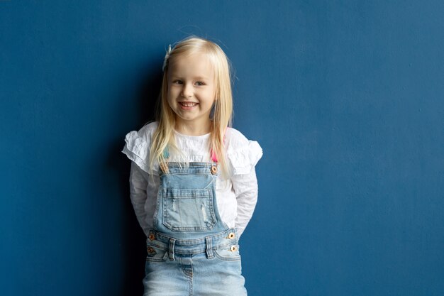 Gelukkig peuter mooi meisje die met blond haar terwijl het stellen op blauwe muurachtergrond glimlachen