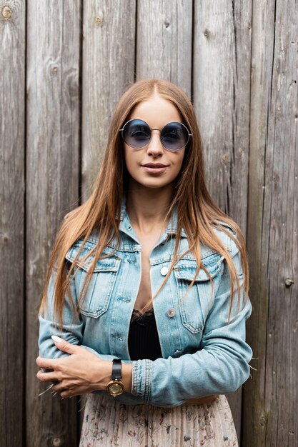 Gelukkig portret van een mooie vrouw in modieuze ronde zonnebril in jeansjasje en vintage rok staat in de buurt van een houten muur