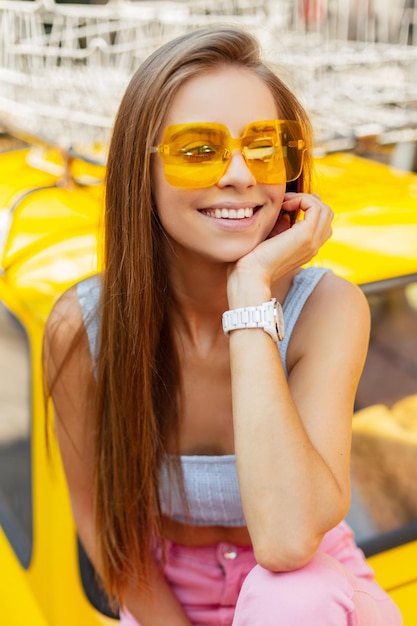 Gelukkig portret van mode jonge mooie vrouw met schattige glimlach in kleurrijke zomerkleding met coole zonnebril zit en rust in de buurt van een gele retro auto