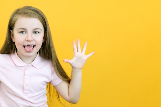 Gelukkig positief glimlachmeisje die vijf vingers tonen die haar tong uitsteken geïsoleerd op gele kleurenachtergrond