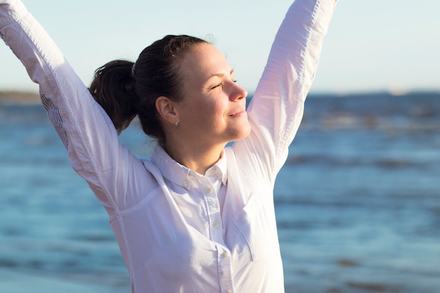 Gelukkig positief mooi meisje, jonge vrouw in wit overhemd genieten van vakantie op zee, strand, stretch, houd haar armen, handen omhoog met gesloten ogen. Goed humeur, ontspanning, geluk concept