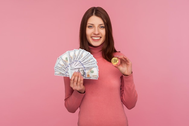 Gelukkig positieve brunette vrouw in roze trui bedrijf in handen bitcoin en dollar biljetten vergelijken crypto valuta met papiergeld reclame Indoor studio opname geïsoleerd op roze achtergrond