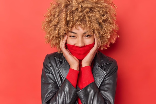 Gelukkig positieve vrouw verbergt mond achter kraag gekleed in leren jas voelt zorgeloos en vreugdevolle poses tegen rode achtergrond heeft ogen vol geluk. Mensen kleding en emoties concept