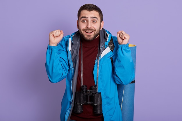 Gelukkig reiziger jonge man in blauwe casual jas met rugzak geïsoleerd op paarse achtergrond. Toerist die op weekenduitstap reist