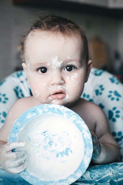 Gelukkig rommelige babyjongen zittend in een kinderstoel en spelen met zijn vuile bord met mooie glimlach