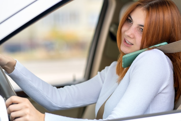 Gelukkig roodharigemeisje die op haar mobiele telefoon achter het wiel spreken die een auto drijven.