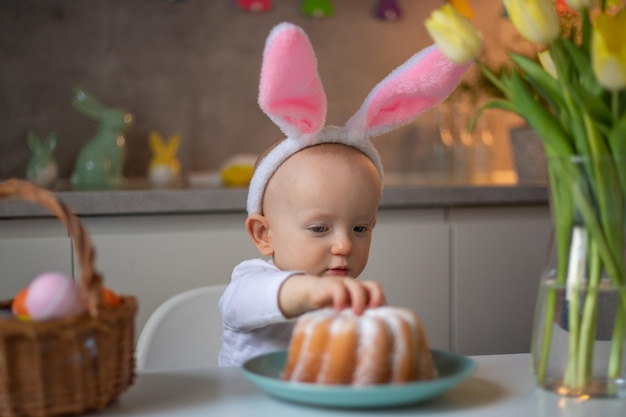 Gelukkig schattig klein babymeisje met konijnenoren op paasdag zittend aan de tafel in de keuken en eet een verjaardagstaart