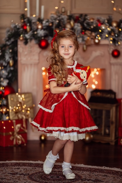 Gelukkig schattig klein blond meisje met lang haar in een rode jurk, wachtend op kerstmis