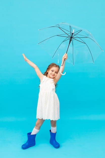 Gelukkig schattig klein meisje in blauwe rubberen laarzen en een katoenen witte jurk met een paraplu op een blauwe achtergrond in de studio en glimlachend en gek rond ruimte voor tekst
