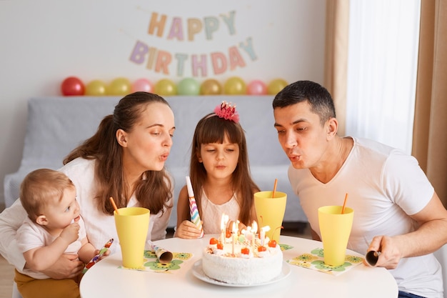 Gelukkig schattig vrouwelijk kind dat een wens doet en kaarsen op de taart gaat blazen tijdens het vieren van de verjaardag met familie, zittend aan tafel met verjaardagsdecoratie op de achtergrond.