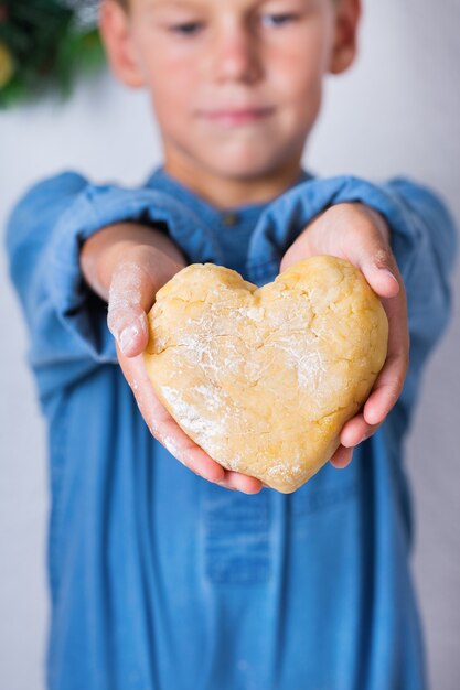 Gelukkig schattige jonge jongen in handen hartvormig stuk zelfgemaakte deeg te houden. Familie tijd in de gezellige keuken. Winteractiviteit in huis. Hulp en huiswerktraining voor kinderen