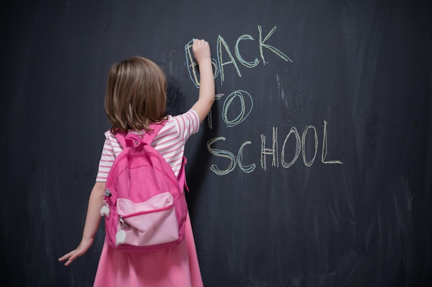 Gelukkig schoolmeisje dat met rugzak terug naar school schrijft op zwart bord